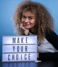 Smiling woman with curly hair beside 'Make Your Choice' sign, promoting voting.